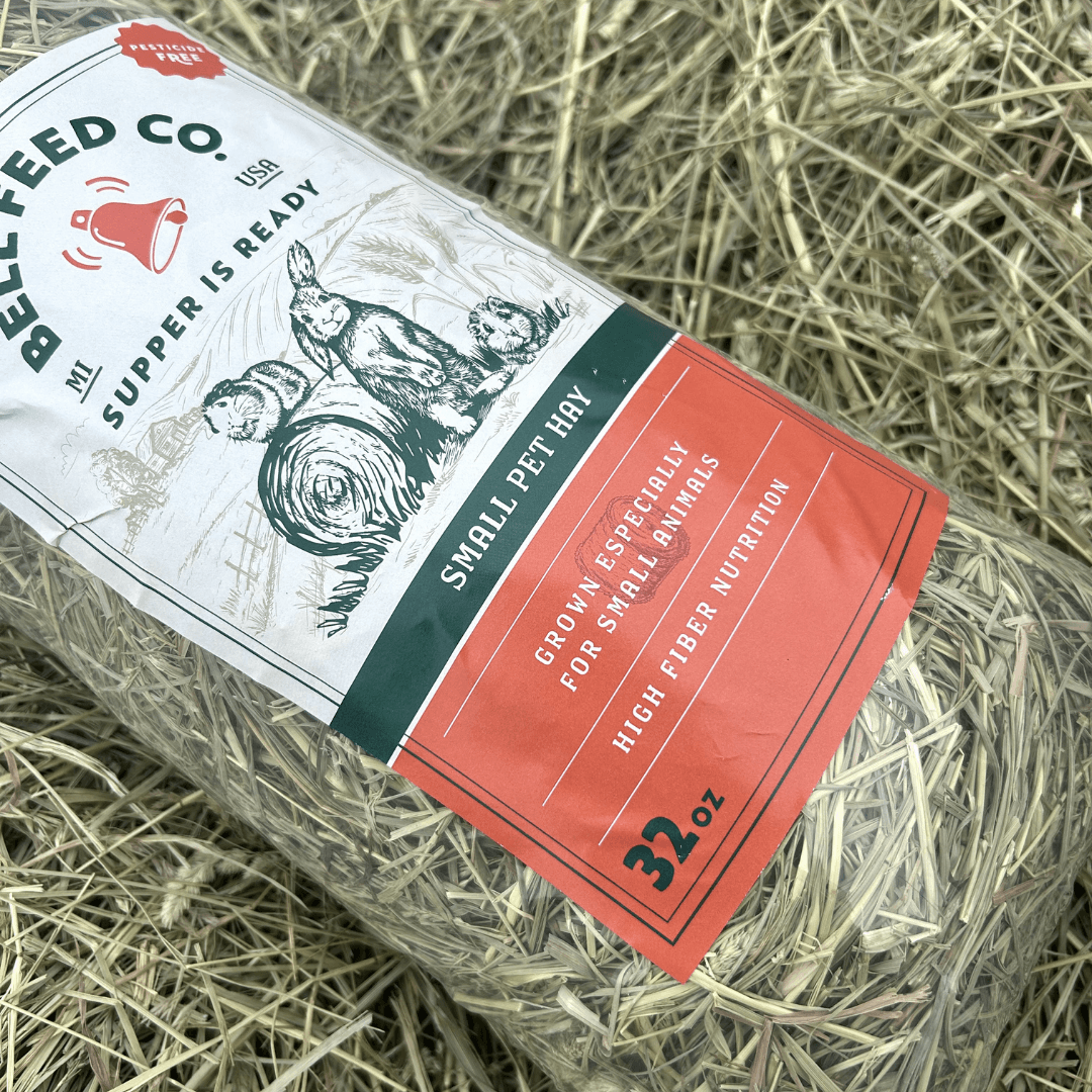 A roll of Bellfeed Co. Small Pet Hay wrapped in a white, dark green, and coral orange label, resting on a bed of hay.